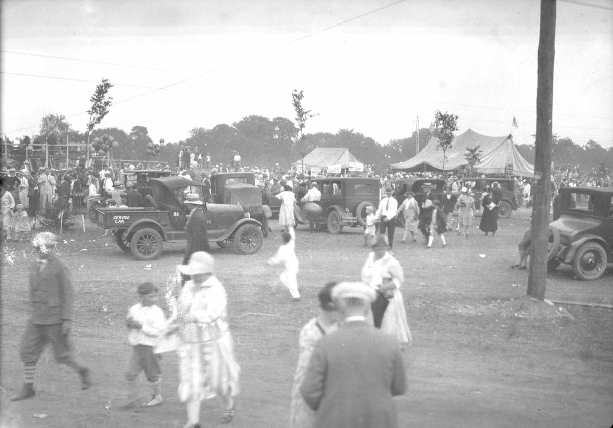 A crowd of people walk through an open field. Some sit in old "model T" style cars.