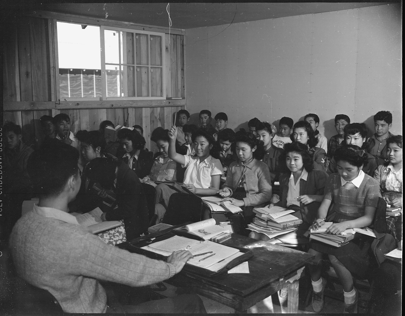 Bottom left we see the back of a man with his hand resting on an open notebook on desk. In front of him, facing the camera are four rows of students of Japanese ancestry with piles of books on their laps. One young woman is raising her hand and smiling.
