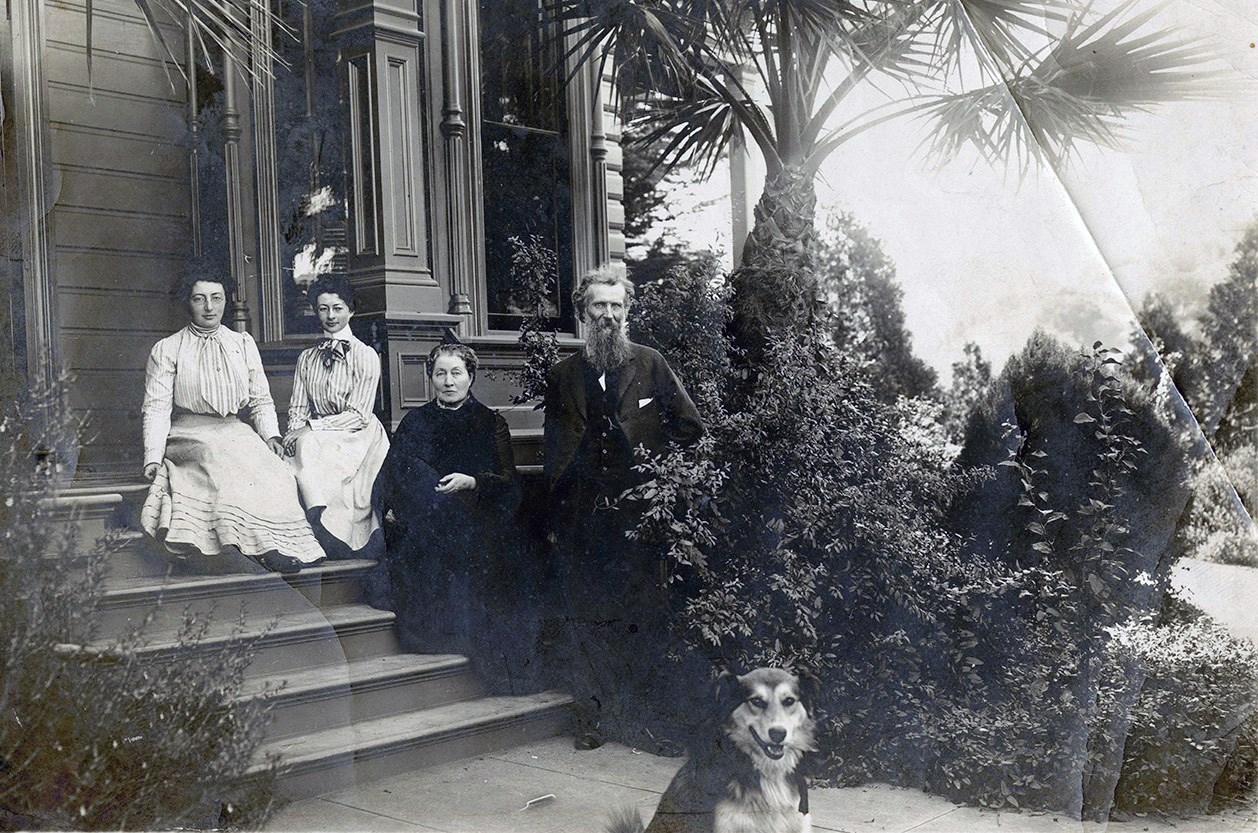 Old fashioned portrait of family sitting on porch, two young women in high-necked dresses, older woman in black, and man in suit standing. Dog sits in foreground