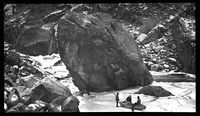 Three men and a boat, on iced over river Black and white image of three men sitting on a wooden boat. The boat is on a section of river iced over. Large boulders surround the river.