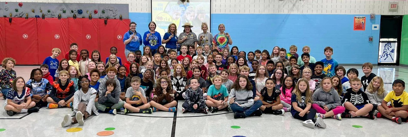 Multiple classes of elementary school students with staff and a park ranger in a school gym.