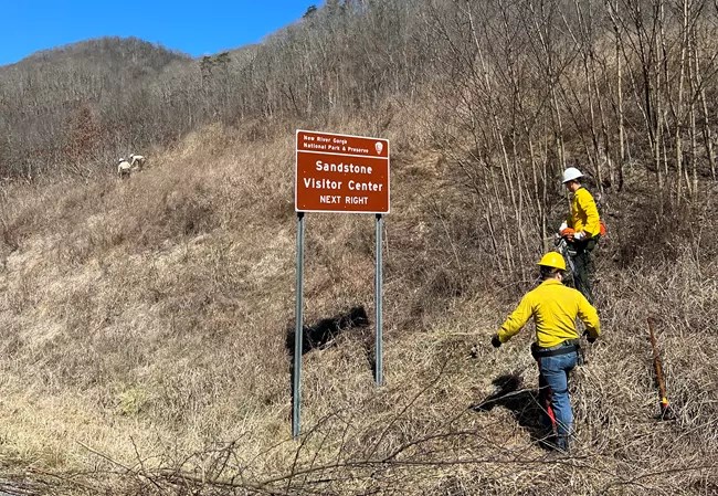 Veterans wildland fire crew at New River Gorge National Park & Preserve Two people in wildland fire gear stand near a sign which reads Sandstone Visitor Center next right.