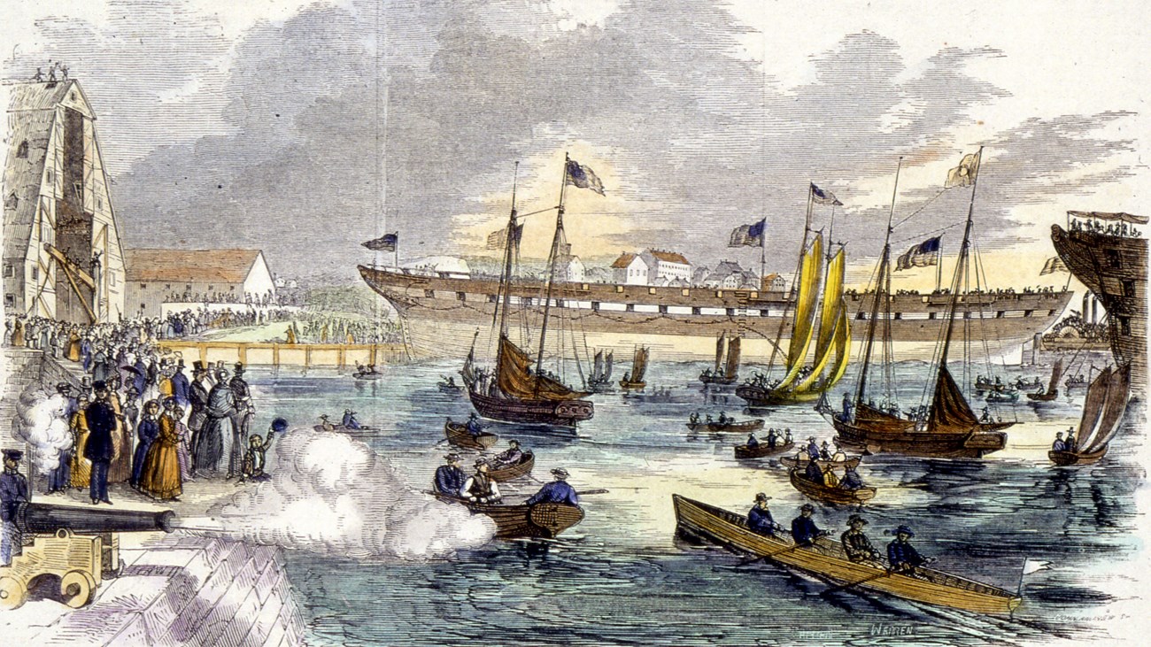 Hull of a ship launching into a harbor as crowds watch on piers and a swarm of boats. A cannon fires in celebration.