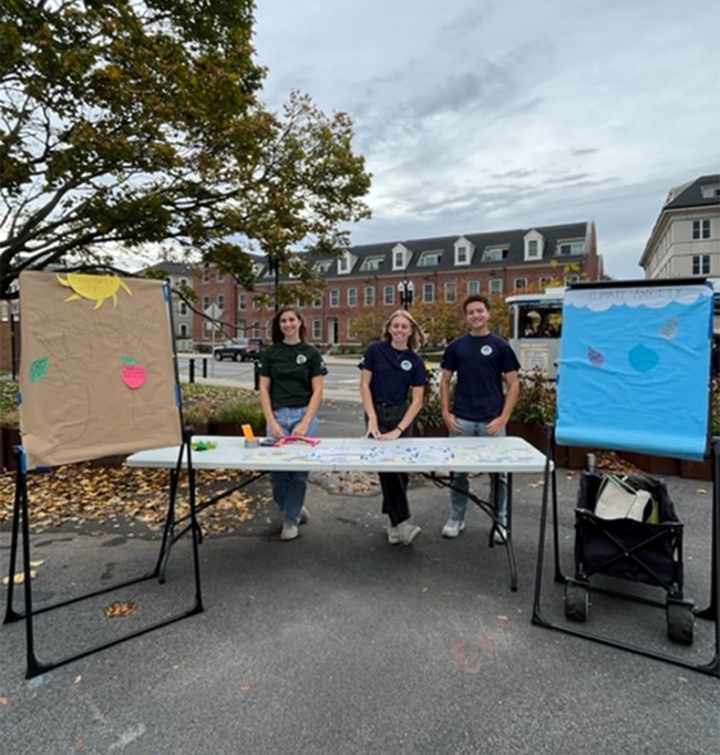 Three people working at a tabling event, smiling towards the camera. They are outside and have a poster on each side of the table displaying climate change haikus.