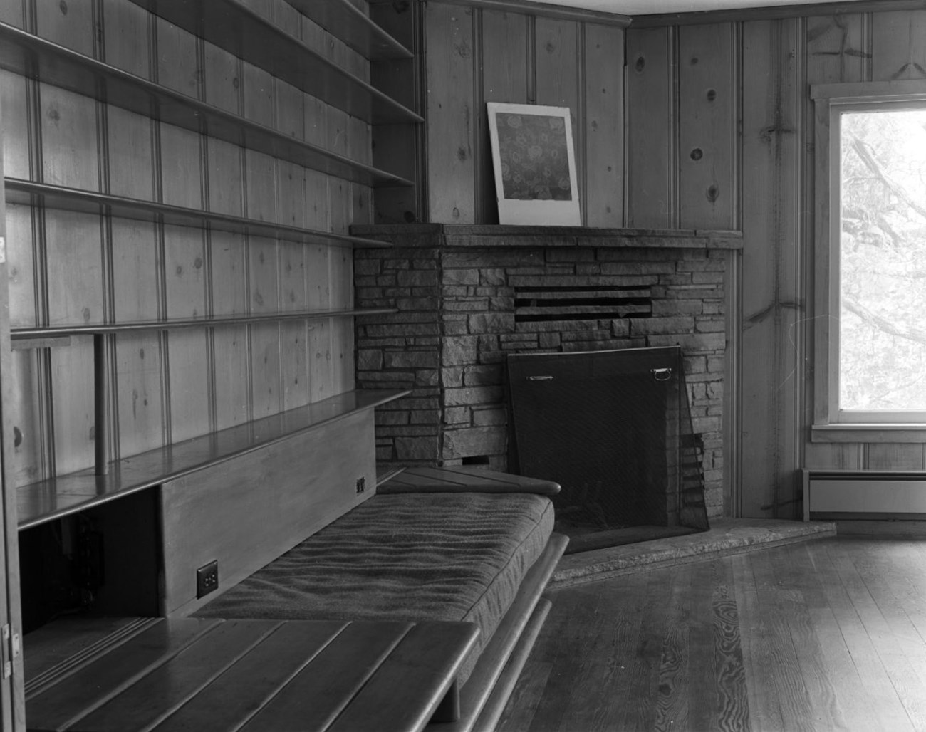 Living room with stone hearth. Wood paneling covers walls, low bench abuts bookshelves on left of fireplace and window to its right looks out into forest.