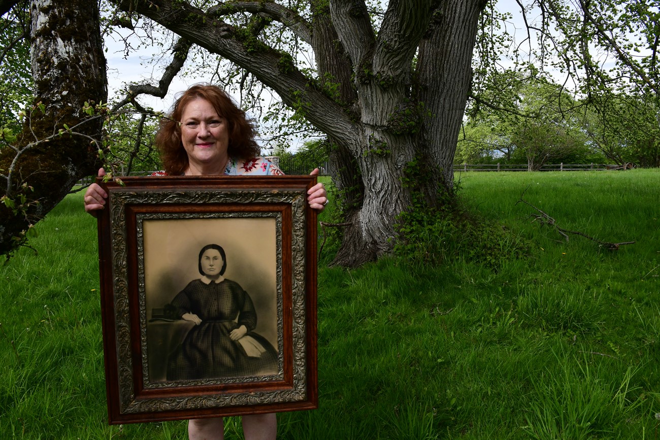 Woman standing in front of tree holding old portrait of another woman