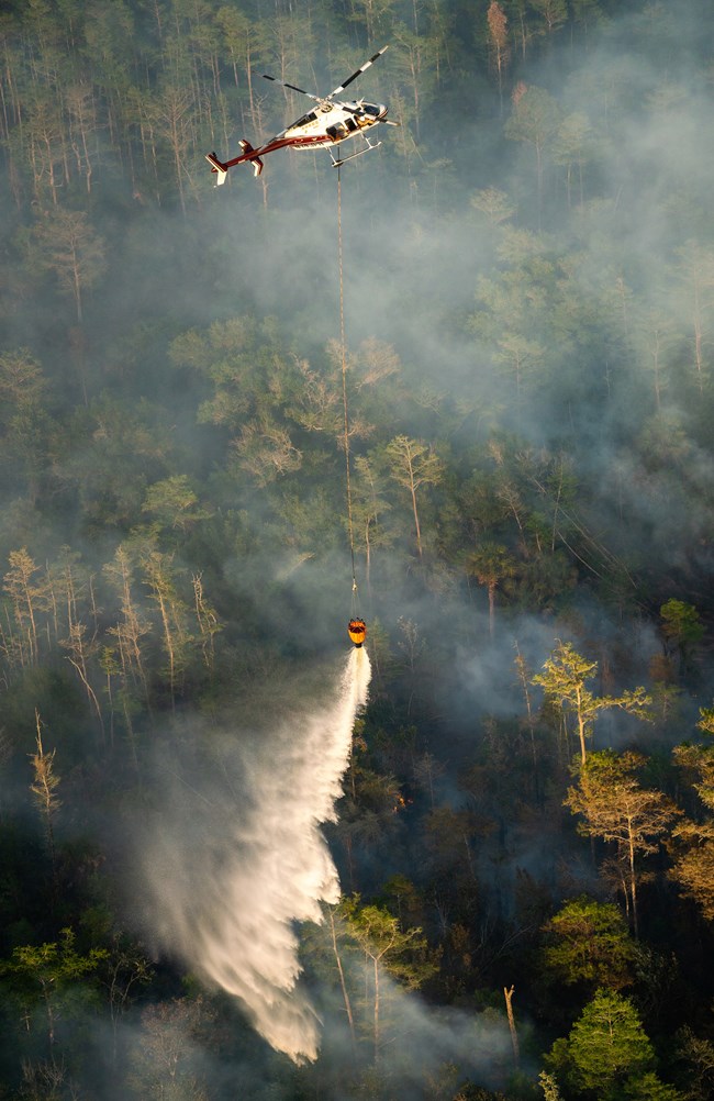 A helicopter drops water from a bucket on a forest fire.