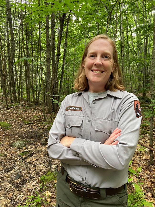 a woman in uniform crosses her arms and smiles at the camera with trees surrounding her
