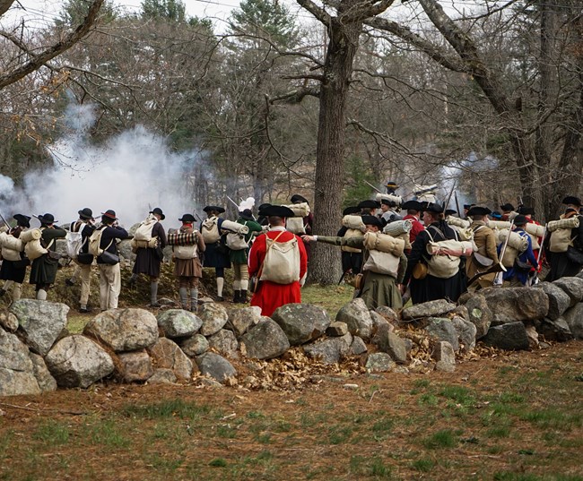 A firing line of Revolutionary War militiamen shown from behind firing their muskets