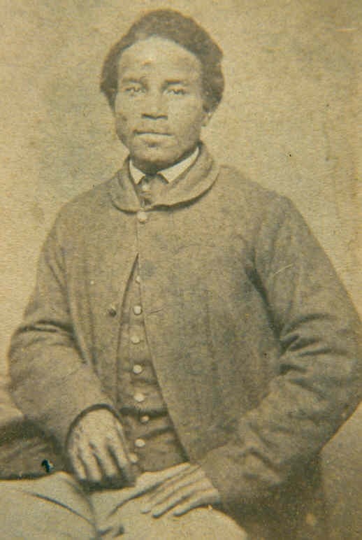 Portrait of an African American man sitting down wearing a coat over a collared shirt and waistcoat.