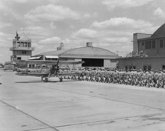 Black and white photo of an airplane on an air strip with soldiers lined up on the right