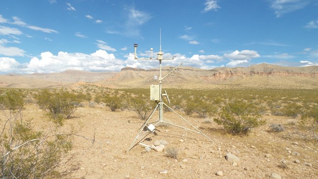Weather monitoring station amidst creosote shrubland slopes of hills and mesas in background.