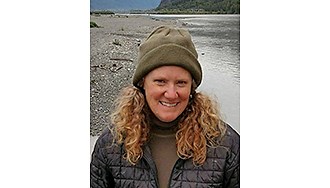 Portrait of Chambers in an olive green beanie and a black puffy jacket by a river, smiling for the camera.