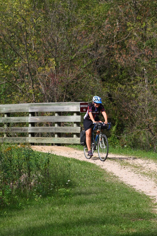 A biker on a dirt trail. A wooden bridge with a brown sign reading "bridge" is behind her on the trail.