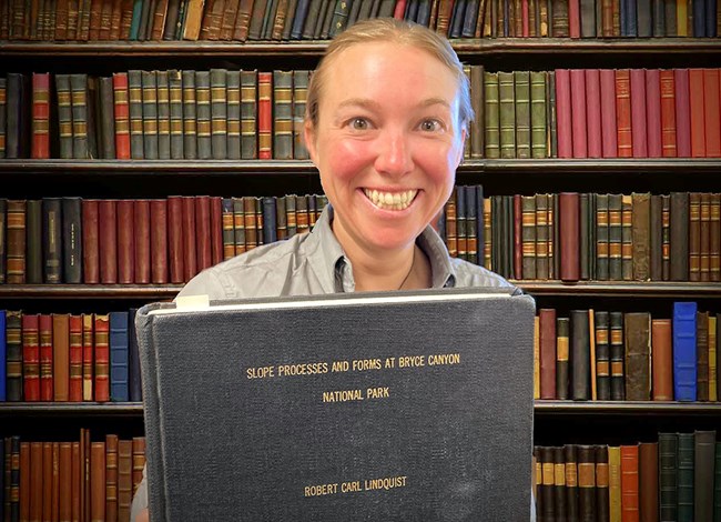Olstad holding Lindquist's dissertation A woman holds a large, gray, old-looking hardcover book in front of a wall of other old books.