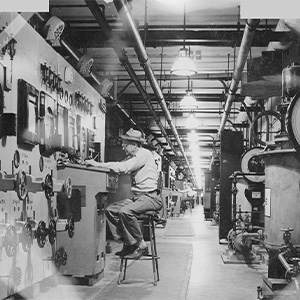  A man sits on a tall stool along the left-hand side of a long hallway, adjusting a dial with his left hand. The walls are covered with dials, levers, and meters.