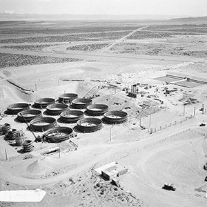 A black and white aerial photo of a desert landscape dotted with industrial buildings and twelve large black circular structures.