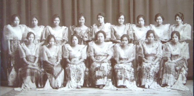 Black and white photo of two rows of Filipino women. The front row is seated; the back row is standing. They are all looking towards the camera. All women are wearing long patterned shirts and tops.