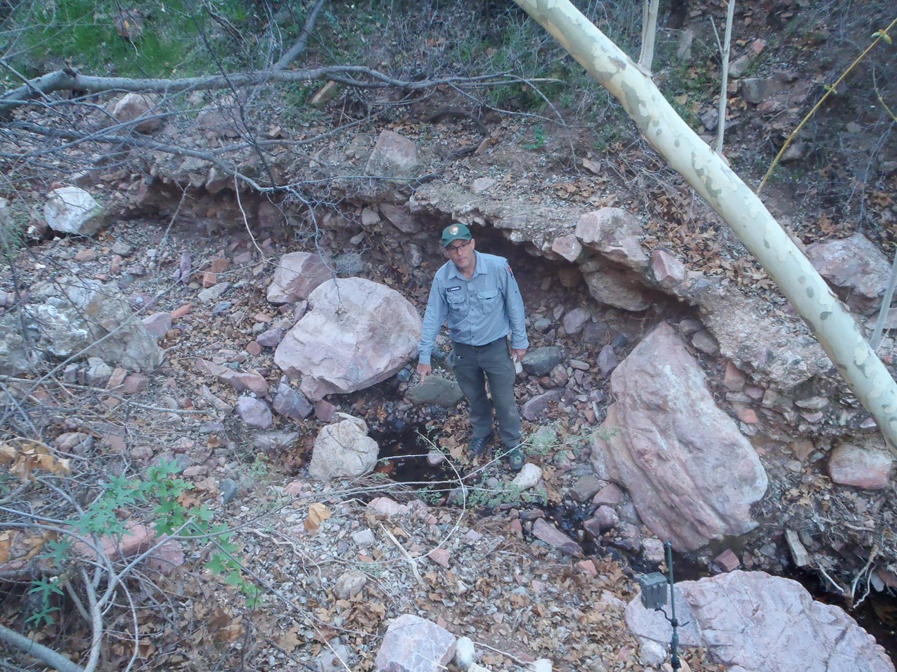 Cave Canyon Spring A man stands next to a small pool that drains in a narrow path down a slope. He points to the center of the pool for the camera.