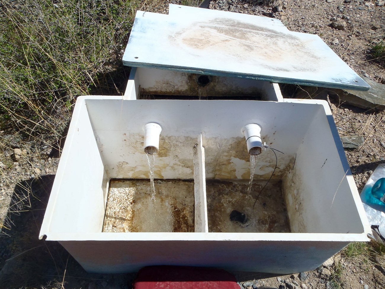 A white box, lid removed, has two pipes inputting water into two equal divisions of the container. Water inside is shallow.