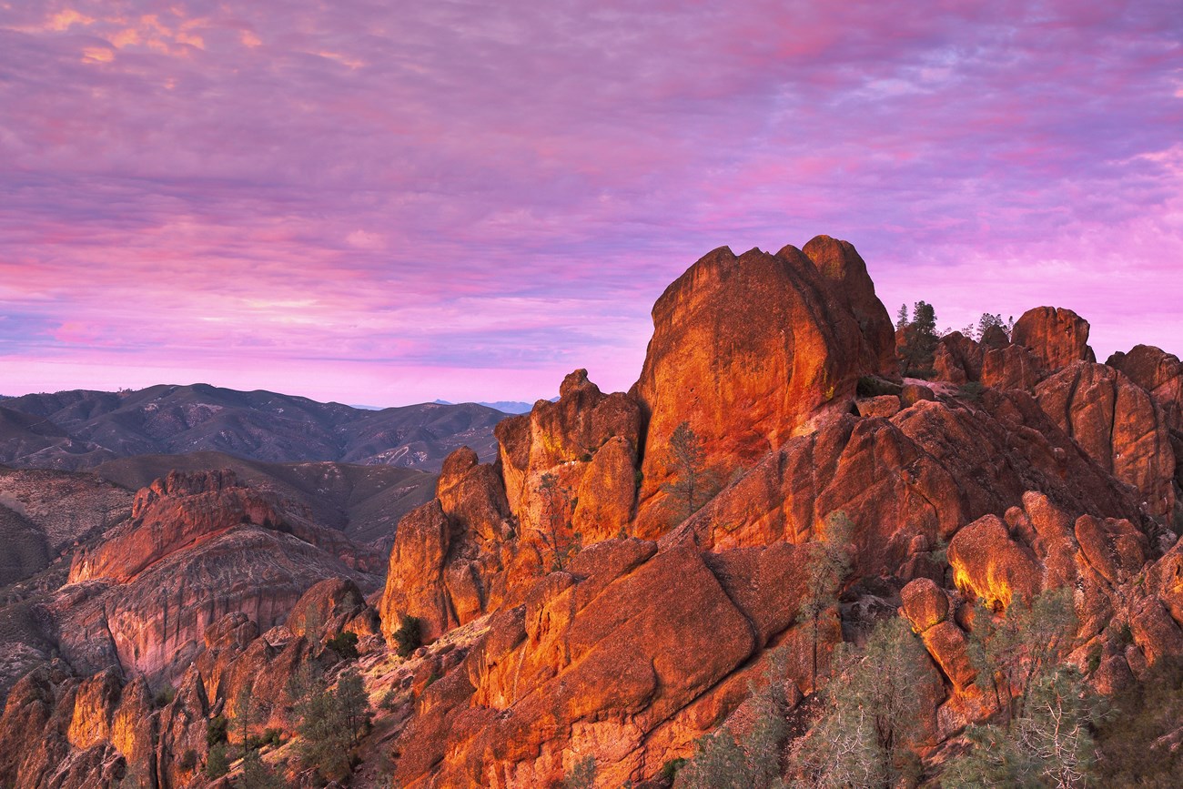 Dramatic rock spires awash in orange glow of sunset, contrasting against purple sky over mountains in distance.