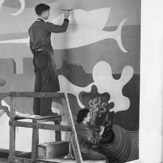Black and white photo of man and woman on scaffold, painting mural.