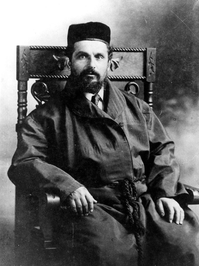 Black-and-white photograph of a bearded man seated in a wood chair with decorative carvings. He is wearing a Bukharian kippah and a dark robe with a tassel belt, worn over a shirt and tie.