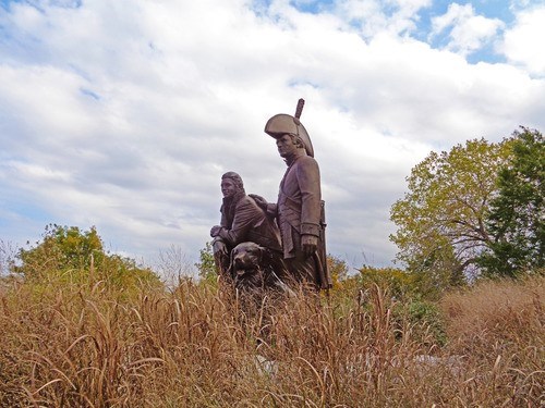 A statue of Lewis, Clark and Lewis’ dog Seaman. Tall, brown grass is in the foreground, and trees and a partially clouded sky are in the background.