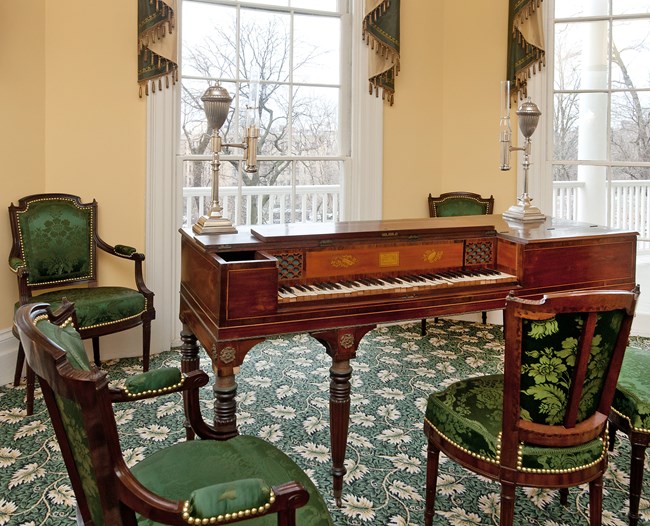 A piano is surrounded by ornate mahogany chairs upholstered in green silk.