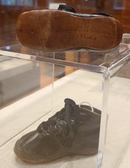 Two high-top black leather baby shoes with shoelaces tied in a bow. On the brown soles there is “Parmly Billings’ First Shoes” in cursive.