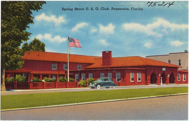 Colorful postcard of a single story red building with a green lawn, an American flag flying on a pole and a blue sky in the background