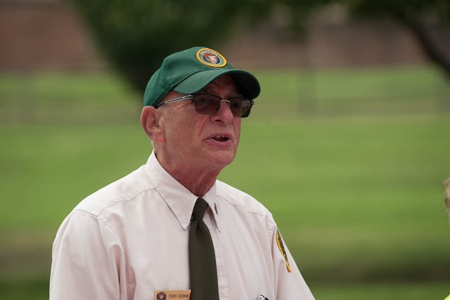 Elderly white male person, close up of his face as he speaks to other people. Background is grass with trees. He is wearing a white shirt with a black tie, and a green baseball cap that has the NPS volunteer logo.