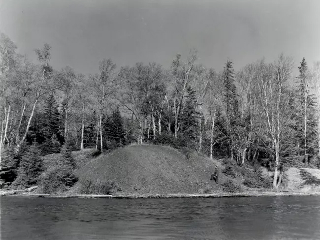 Black and white photo. In the middle-ground, surrounded by birch and conifer trees, is a pile of dark rock on the shoreline and a man climbing it. In the foreground is calm water.