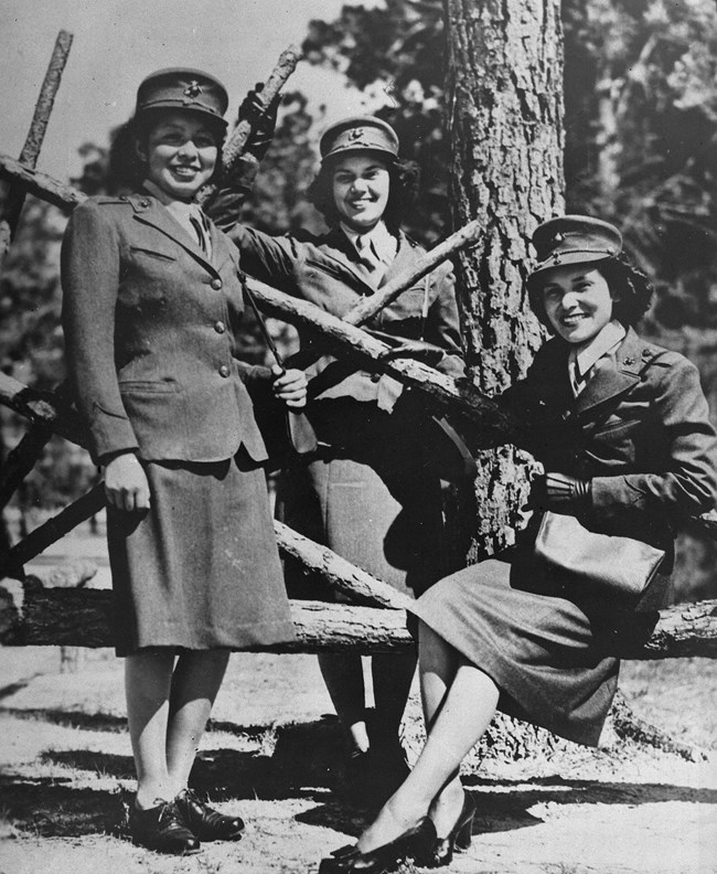 Three young women in military uniform smile at the camera; two stand and one is seated on a log fence