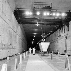 A black and white photo of a cavernous building. A pulley system is moving equipment through the building while two workers look on.