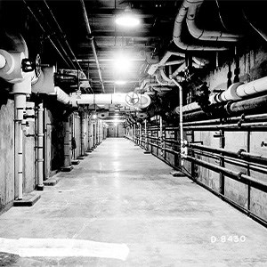 A black and white photo of a large concrete hallway with numerous pipes of varying sizes running along the walls and ceiling.
