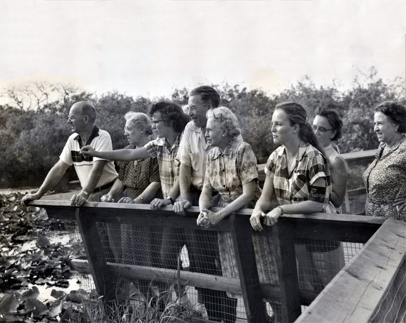 Eight people stand on a boardwalk look out over water. A girl points towards something off camera. One girl wears a ranger aide armband.