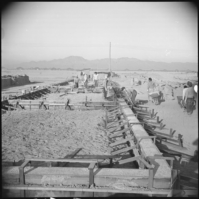 Black and white photo of construction site. blurry figures with wheelbarrows on left. Flagpole in background.