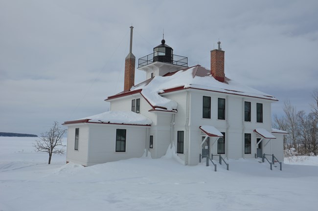 A white lighthouse station with red trim covered in snow.