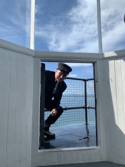 A man dressed in a black lighhouse keeper uniform crouched at the top of the small door leading out to the top of the lighthouse platform.