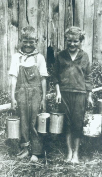 A black and white photo of two children, a boy and girl, standing  barefoot in front of a wooden fence holding buckets of berries in each hand.