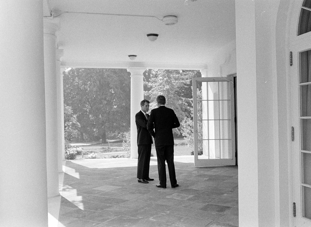A black and white photo of RFK and JFK outside the Oval Office.  JFK's back is turned to the camera and RFK looks pensive.