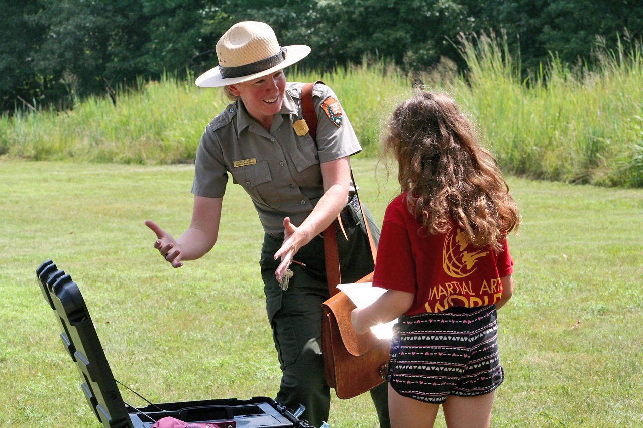Park ranger in the field Woman outside in a green and gray uniform talking with a young girl and gesturing towards items in a black trunk.