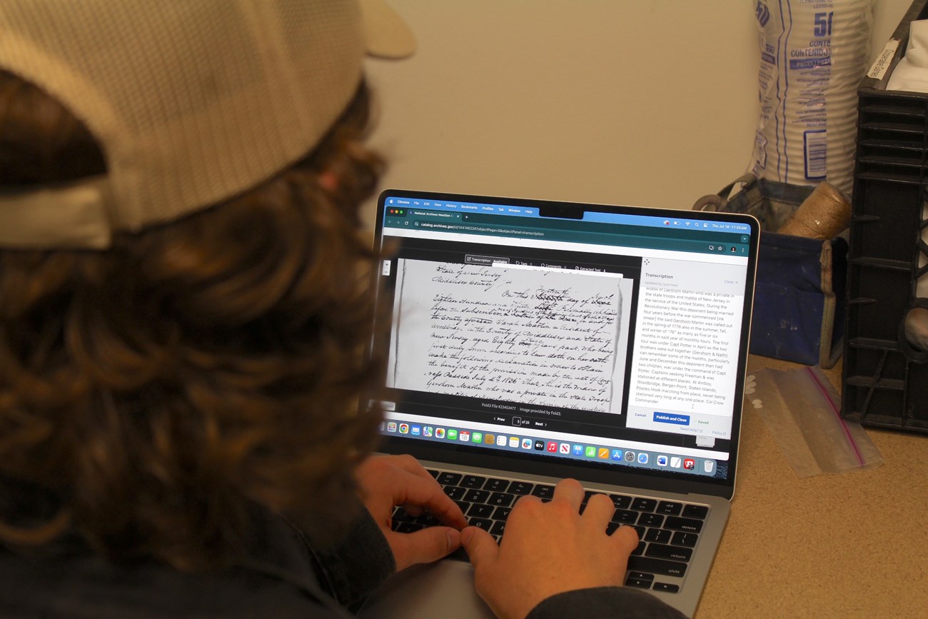 A computer screen shows a historic document written in cursive as a person in a white cap types a transcription.