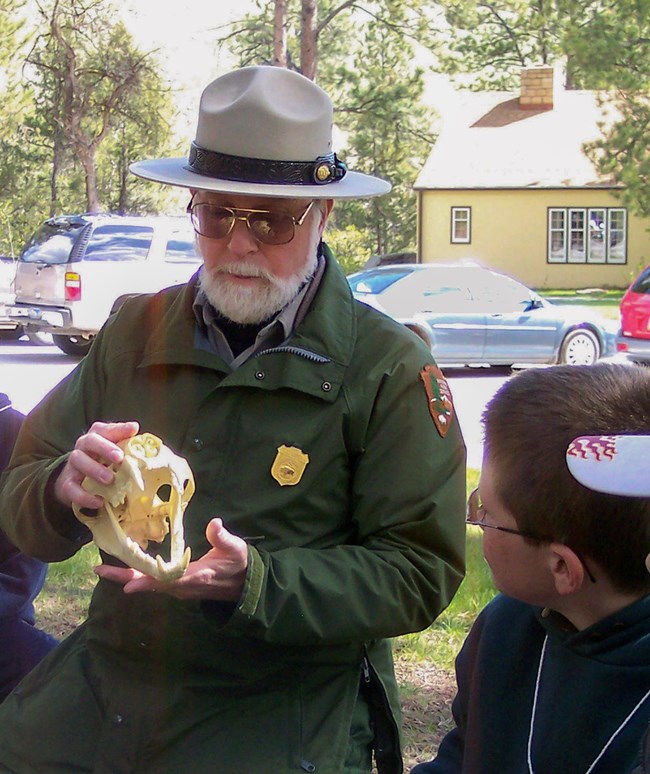 A Cauciasan man with a white beard wearing a green coat and ranger hat sits on a lawn holding a plastic model of an animal skull. A young student next to him dressed in dark clothing looks at the skull.