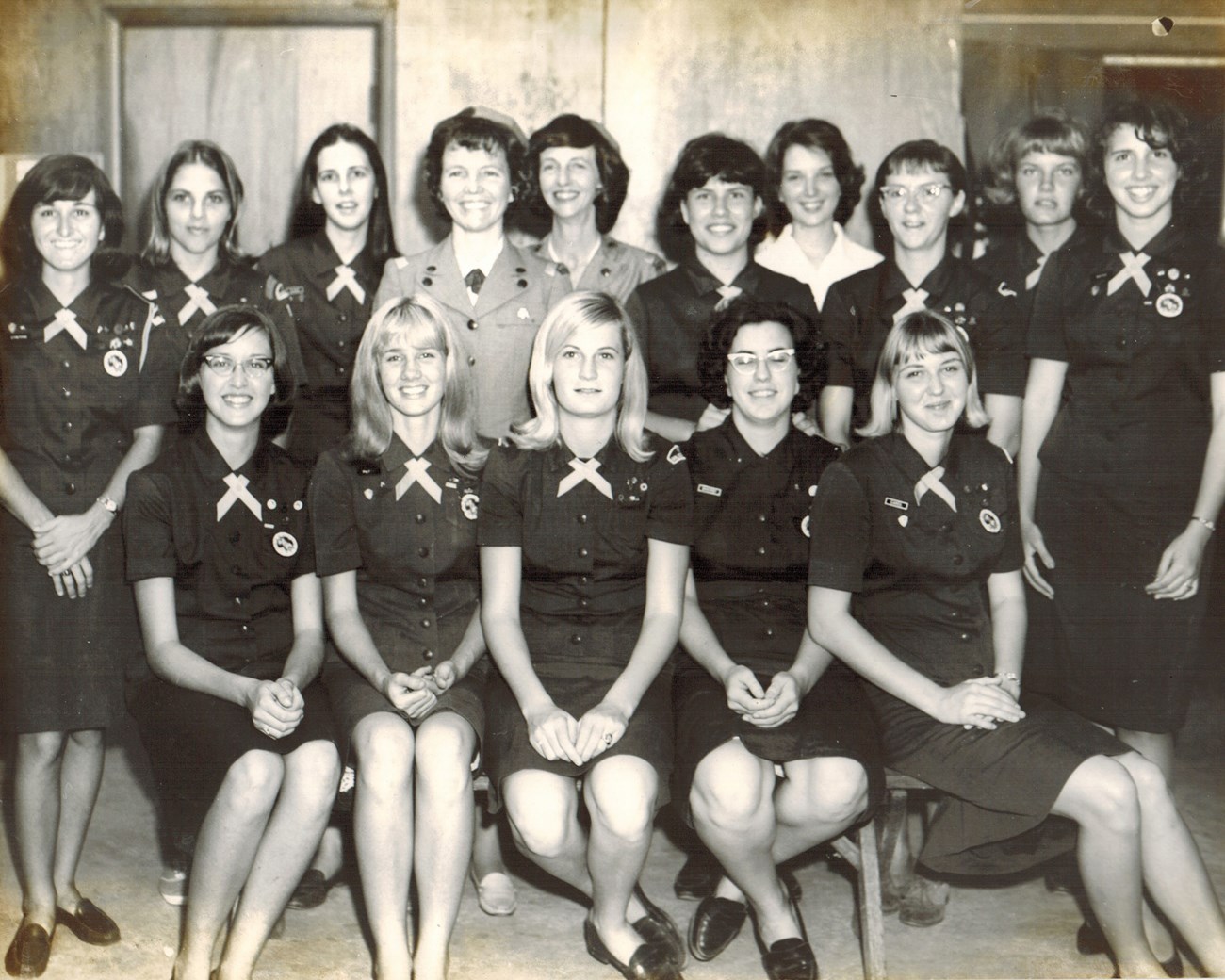 Thirteen girls and two troop leaders pose in their Girl Scout uniforms of skirt, shirt, and cross tie. Only the leaders wear hats and jackets.