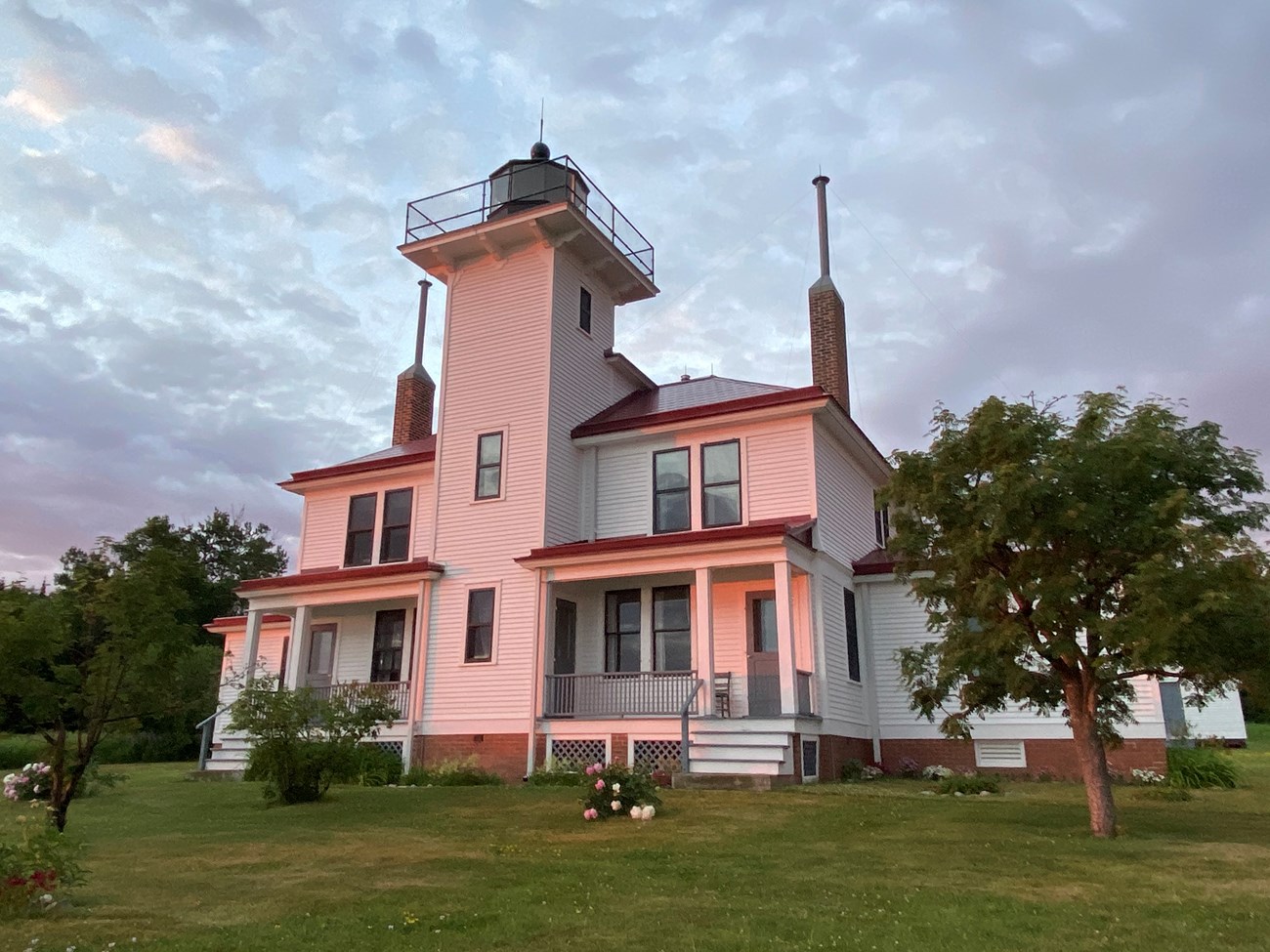 A white lighthouse station with red trim and green lawn glows pink at sunset.