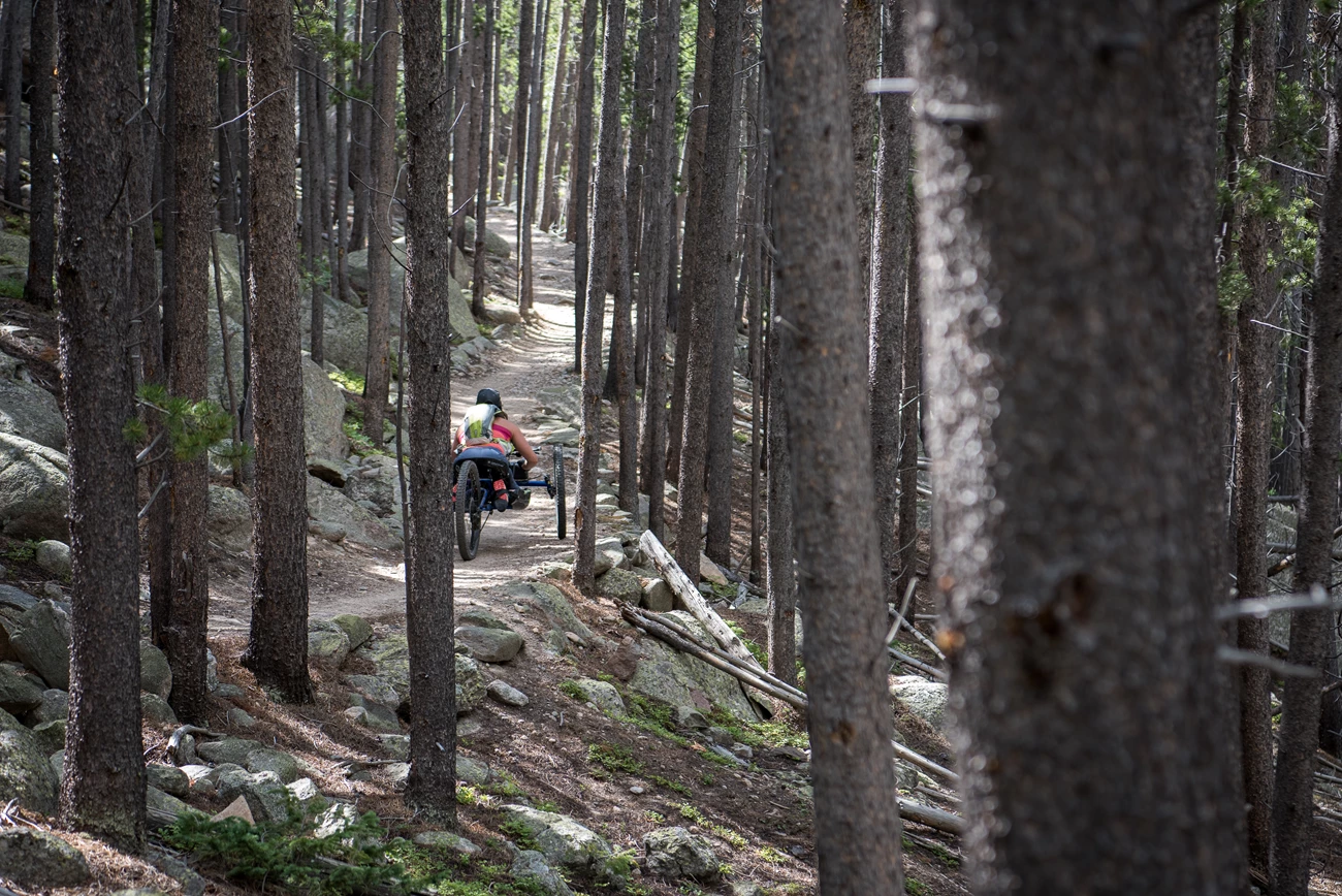 Lawn Lake a woman uses an adaptive wheelchair on a dirt trail through the trees