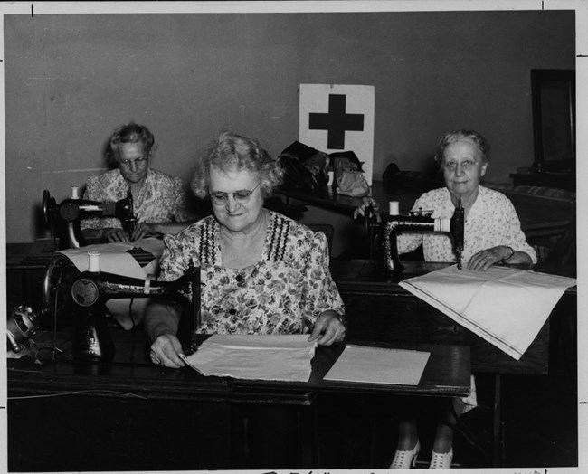 Three senior-age women use sewing machines to work on white square pieces of cloth at sewing tables. The Red Cross logo is hanging on the wall behind them.