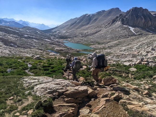 Three people carrying backpacks hike down a rocky slope toward a mountain lake.